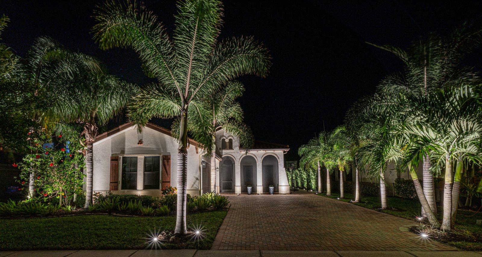 Mediterranean-style Ave Maria home at night with uplit royal palms over a paver driveway